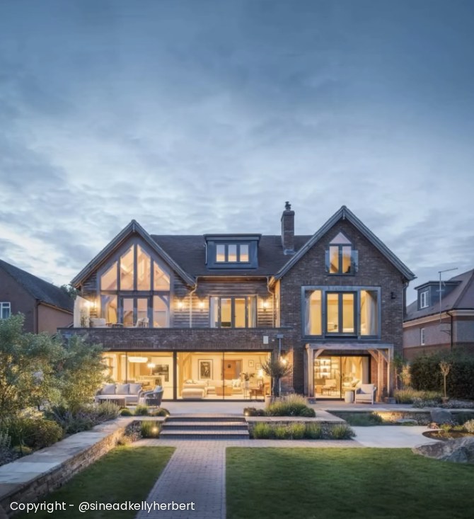Rear of the house at dusk, lit from within, with the oak-framed gable and landscaped garden