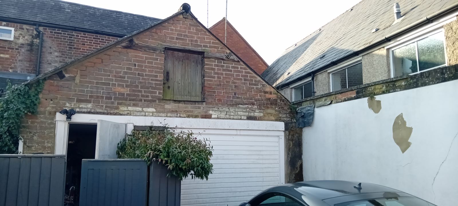 Brick outbuilding gable with a decayed timber loading door and peeling render on the boundary wall, photo 3 of 4